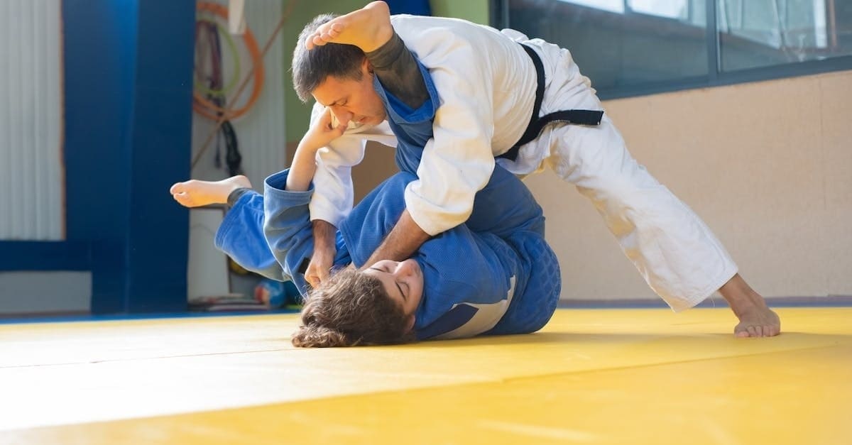 A martial arts instructor in a white gi demonstrating a self defense ground technique, controlling an opponent wearing a blue gi on a yellow mat.